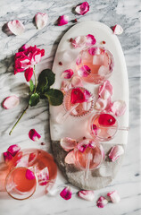 Summer refreshing cold beverage drink. Flat-lay of rose lemonade with ice in glasses and jug and fresh rose flower petals on board over grey marble table background, top view