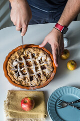 Man cutting traditional apple pie in ceramic baking form, apples and plates on white kitchen.