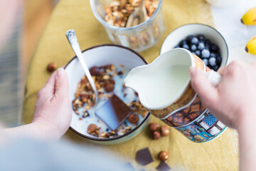 Making breakfast. Pouring milk into bowl with homemade granola with nuts, dark chocolate and bilberries.