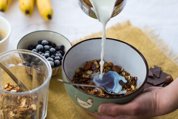 Making breakfast. Pouring milk into bowl with homemade granola with nuts, dark chocolate and bilberries.
