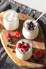Three glass jars with natural homemade yogurt with frozen strawberries, bilberries and jam on wooden board  on white table  .