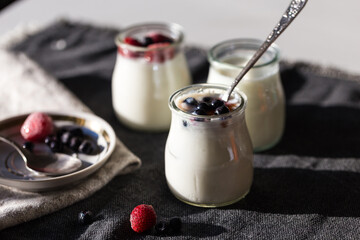 Three glass jars with natural homemade yogurt with frozen strawberries, bilberries and jam on black fabric on white table  .