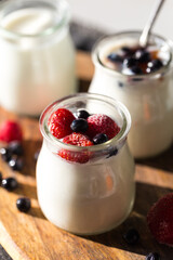 Three glass jars with natural homemade yogurt with frozen strawberries, bilberries and jam on black fabric on white table  .