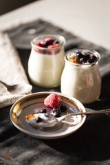 Spoon on plate and two glass jars with natural homemade yogurt with frozen strawberries, bilberries and jam on black fabric background .
