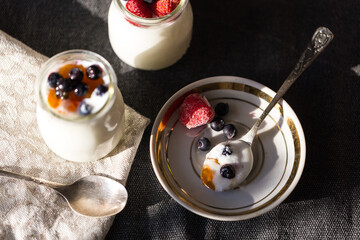 Spoon on plate and two glass jars with natural homemade yogurt with frozen strawberries, bilberries and jam on black fabric background .