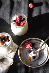 Spoon on plate and two glass jars with natural homemade yogurt with frozen strawberries, bilberries and jam on black fabric background .