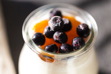  Glass jar with natural homemade yogurt with frozen bilberries and apricot jam on black fabric background .