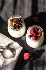 Two glass jars with natural homemade yogurt with frozen strawberries, bilberries and jam on black fabric background .
