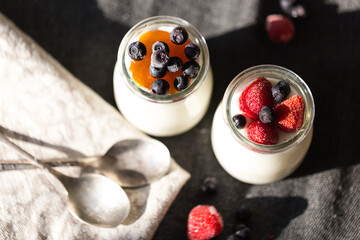 Two glass jars with natural homemade yogurt with frozen strawberries, bilberries and jam on black fabric background .