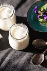 Two glass jars with natural homemade yogurt, spoons, little vase with violets on black fabric on white wooden table.