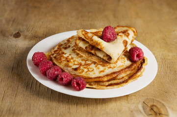 Tasty little thin pancakes with red raspberries on a white plate on a wooden table. Sweet dessert.
