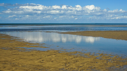 Reflection of clouds and blue sky in the water at low tide with sandy beach in the foreground. Burrum Heads, Queensland, Australia.