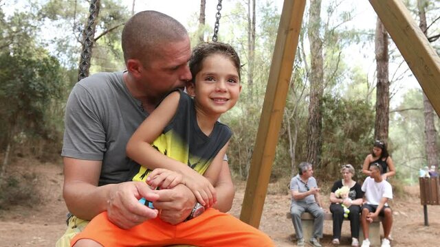Video in 4k di un papa bianco che gioca con il figlio su un altalena in un parco mentre il resto della famiglia alle spalle si rilassa