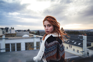 Photo of beautiful girl with long red hairs wearing jacket and looking to camera. Female model standing on flat roof of building.
