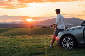Young man resting during long solo auto trip sitting on his luxury car bonnet and enjoying the sunset sky colors. Traveling by car concept image.