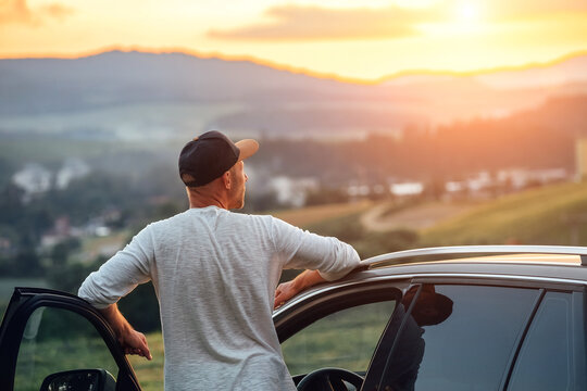 Young man having the long auto trip break. He stoped new car got out of the vehicle and enjoying the sunset orange-pink sky colors.  Traveling by car by the empty nature vastness concept image.