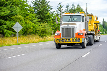 Bonnet powerful big rig semi truck with orange fenders ang oversize load sign on the bumper transporting heavy-duty tractor on the step down semi trailer running on the straight multiline highway road