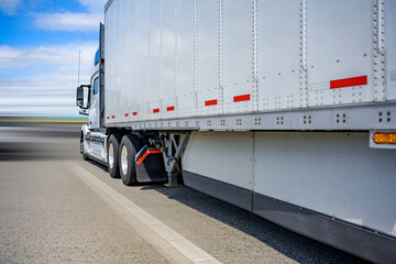 Classic white powerful big rig semi truck transporting cargo in dry van semi trailer with aerodynamic skirt running on the wide straight road to point of delivery