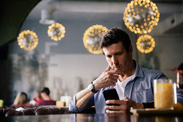 Handsome masculine modern businessman in blue shirt sitting in cafe in thoughtful pose looking at smartphone
