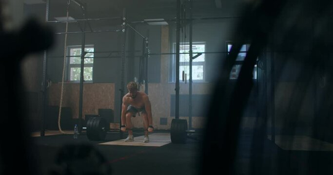 Wide shot of young Caucasian extreme weightlifting athlete man working out with heavy barbell in large hardcore gym hall.