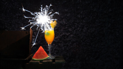 
Orange cocktail in a stemmed glass decorated with a heart-shaped fruit and a firework candle, accompanied by a piece of watermelon