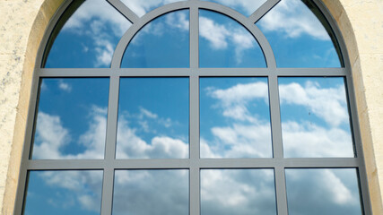 
Close-up on a pretty arched window, the blue sky and its cottony clouds, reflecting on the windows