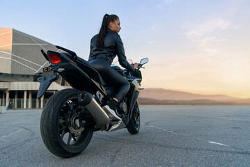 Fototapeta na wymiar Attractive girl with long hair in black leather jacket and pants on outdoors parking with stylish sports motorcycle at sunset.