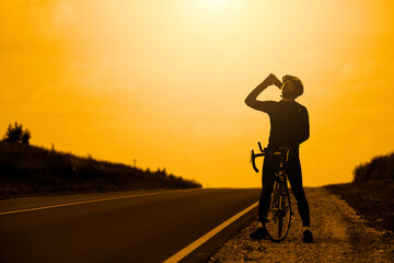 Fototapeta na wymiar The silhouette of a professional cyclist in sportswear wearing a helmet standing with a Bicycle on an open road against the background of the sunset sky