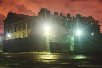 A night view of protected creepy penitentiary prison building facility, asylum exterior with barded wire on walls, red night sky, concept of scary house with maximum security