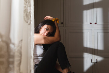 Hispanic young woman sitting in her room with sunlight on her face, relaxed woman thinking looking at the camera, young woman sitting near her window with white curtains