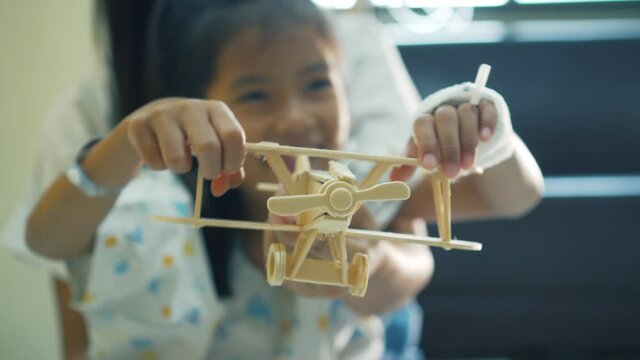 Sick asian little girl playing toy wooden airplane with her mother together while stay in the hospital bed. Therapy for patients to relax. Recovering little girl. 