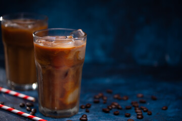 Cold coffee with ice cubes and milk on a background with coffee grains and straws, dark blue background, summer, cool drinks with space
