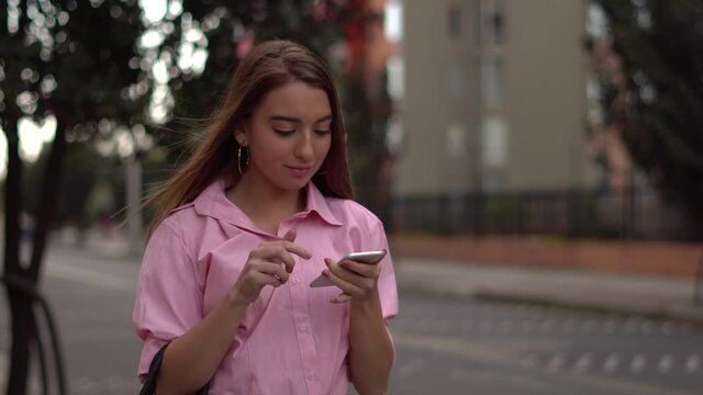 Mujer joven cauc&aacute;sica camina por la calle mientras mira el celular. Revisa las redes sociales, mirando likes y comentarios.
