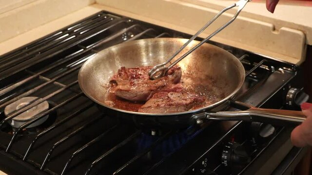 Butterflied lion steak frying in a skillet on a stove top; woman flipping the steak