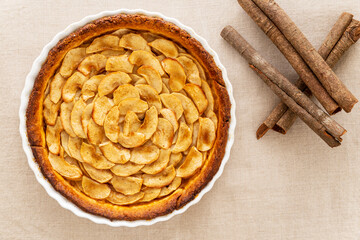 Homemade baked French apple tart, an open faced apple pie, in a baking white ceramic dish aside cinnamon sticks, all on a natural linen table cloth. Flat lay, top view.