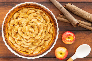 Homemade baked French apple tart, an open faced apple pie, in a baking white ceramic dish aside Gala red apples, cinnamon sticks and white cooking spoon, all on a vintage wood background. Flat lay