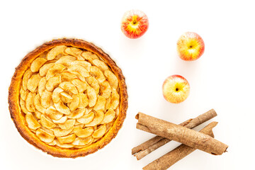 Homemade baked French apple tart, an open faced apple pie, in a baking white ceramic dish aside Gala apples and cinnamon sticks on a white background. Flat lay, top view.