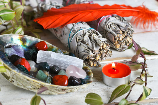 An image of several tumbled healing crystals in an abalone shell with white sage bundles and sacred feather. 