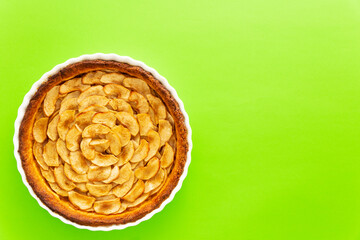 Homemade baked French apple tart, an open faced apple pie, in a baking white ceramic dish on a green background. Flat lay, top view with copy space.