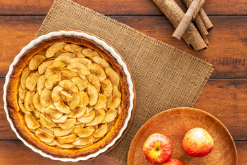 Fototapeta na wymiar Homemade baked French apple tart, an open faced apple pie, in a baking white ceramic dish aside Gala apples in a wooden plate and cinnamon sticks, all on a jute mat on a vintage wood table. Flat lay