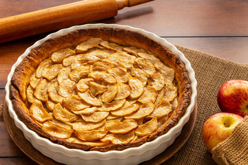 Homemade baked French apple tart, an open faced apple pie, in a baking white ceramic dish aside Gala apples in a jute bag on a vintage wood table.