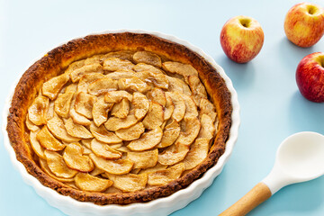 Homemade baked French apple tart, an open faced apple pie, in a baking white ceramic dish aside Gala apples and a white cooking spoon on a light blue background. Side view.