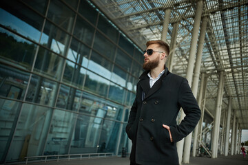 Success. Stylish young bearded man in coat, sunglasses businessman standing near the business center, looking far. With hands pockets, pensive and concentrated. Confident look of