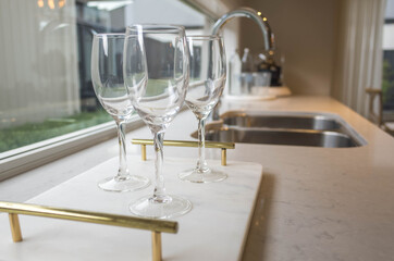 Close up of some wine glasses on marble tray placed on kitchen bench near sink with a blurry view of a kitchen with the neutral color interiors.  Copy space for text.