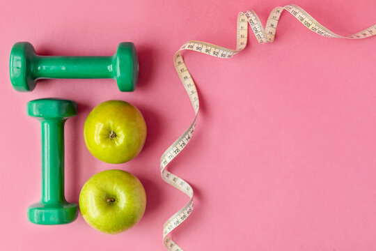 Composition with dumbbells, twisted measuring tape and green apples on a pink background, top view. Concept of diet and healthy lifestyle
