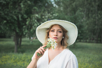 Beautiful girl in a white dress and a big white hat with a bouquet of daisies in her hands poses for the photographer