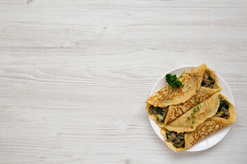 Savory Homemade Mushroom, Spinach and Cheese Crepes on a white plate on a white wooden background, top view. Flat lay, overhead, from above. Copy space.