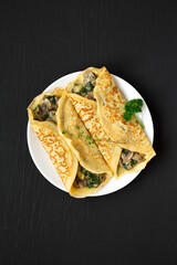 Savory Homemade Mushroom, Spinach and Cheese Crepes on a white plate on a black surface, top view. Flat lay, overhead, from above.