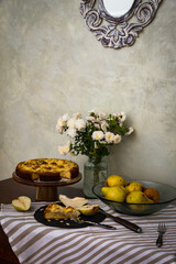 Slice of homemade pear pie and sliced fresh pear on black plate with whole pie and pears on stripped tablecloth .