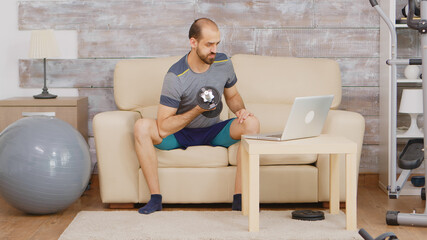 Fototapeta premium Guy training biceps with dumbbell following online training on laptop.